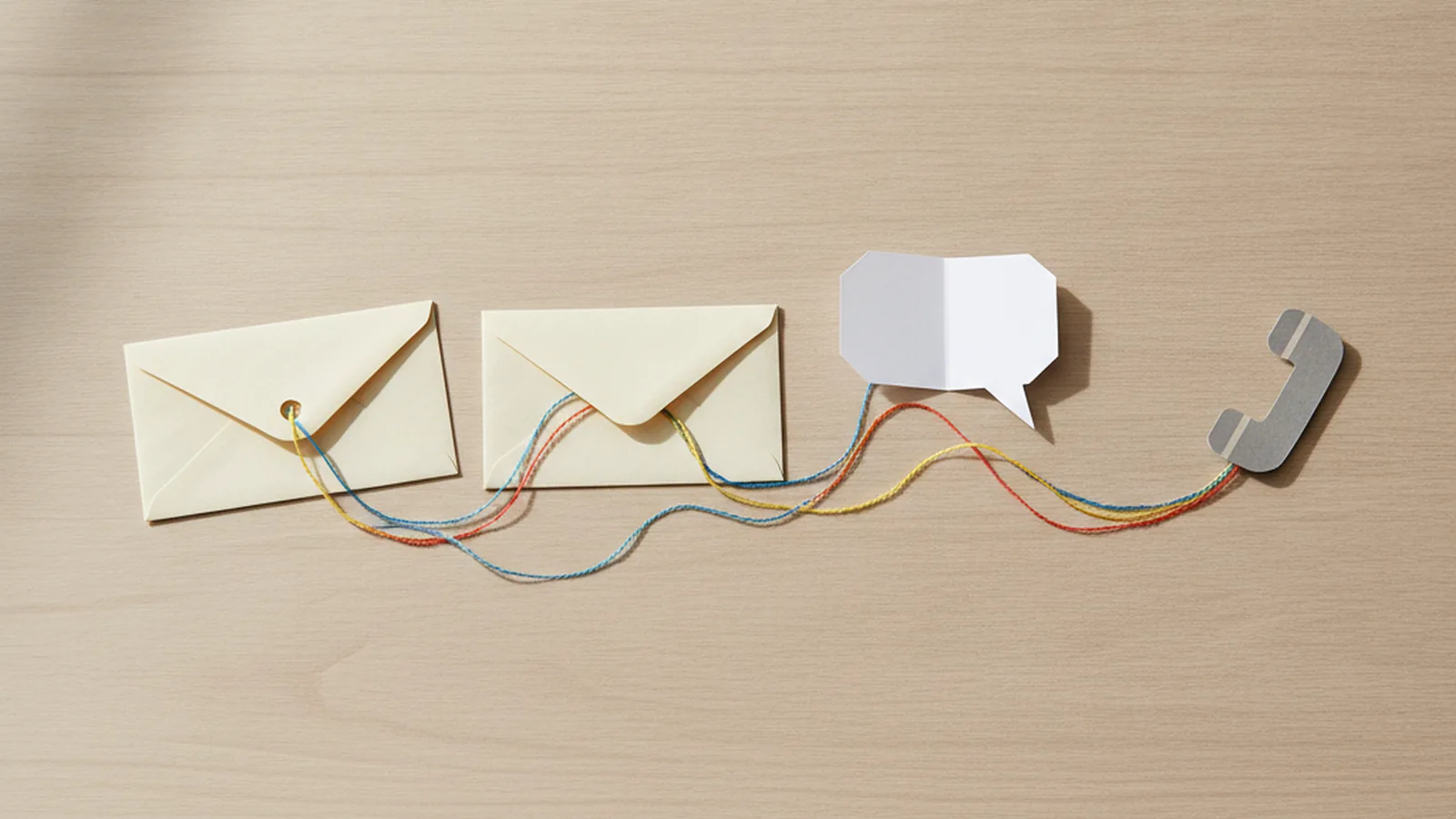 Paper envelopes, a speech bubble and a phone handset on a desk joined by coloured threads